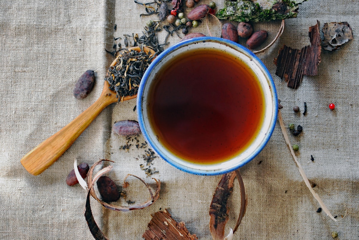 Top-down view of a ceramic bowl of dark tea on rustic linen surrounded by loose leaves and spices
